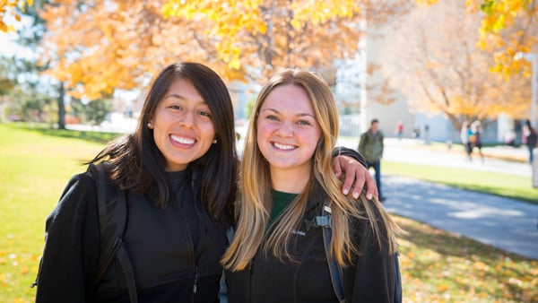 Two female students smiling outdoors on the BYU-Idaho campus in the fall.