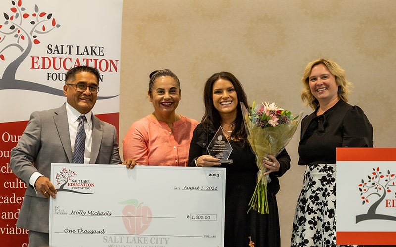 School administrators awarding an over-sized check to a teacher who is holding flowers and a plaque.