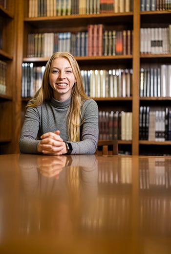 Smiling female seated at table in front of bookcase.